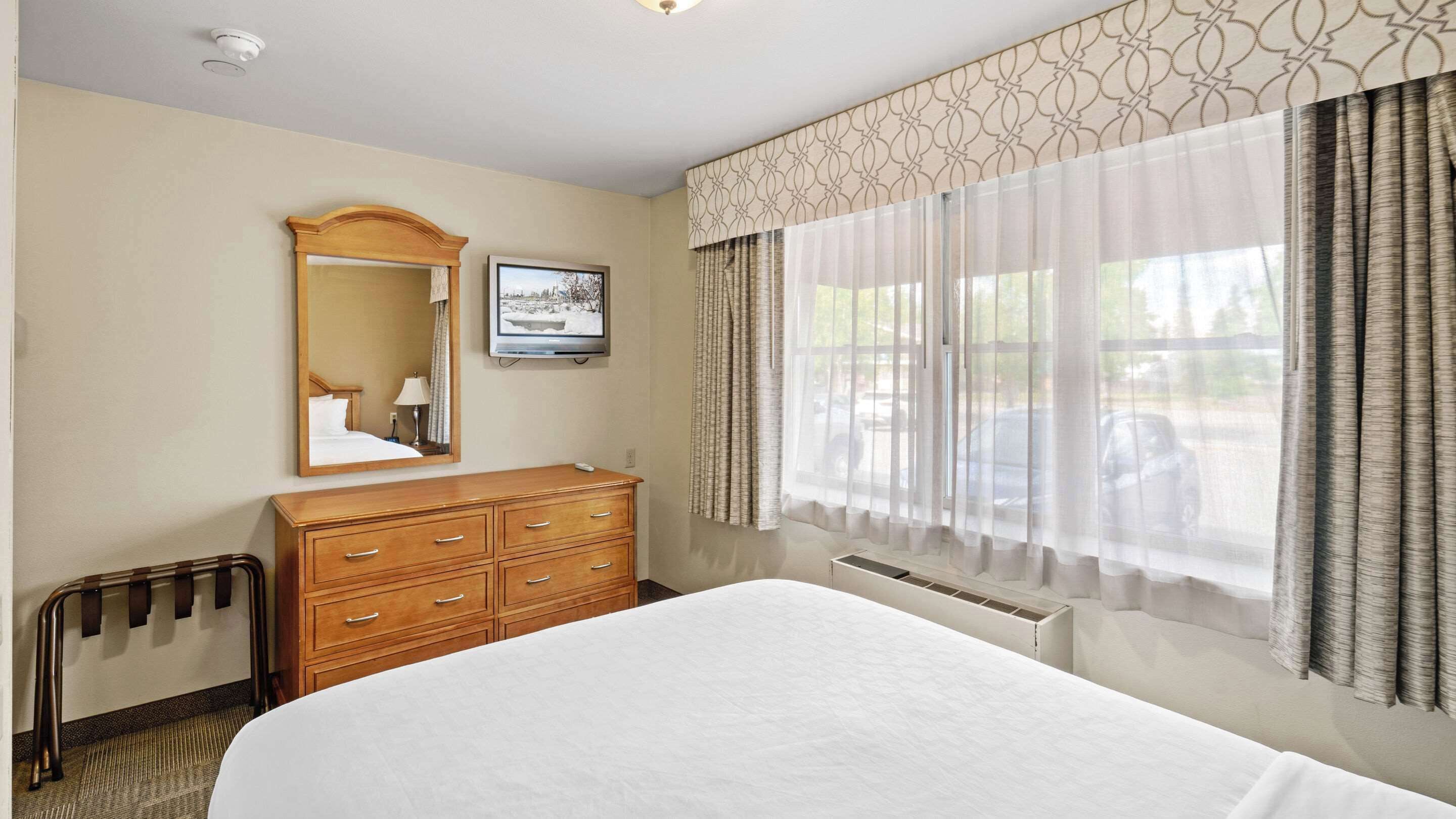 Habitación de hotel con una cama grande, un aparador de madera, un espejo, un televisor y una ventana con cortinas.