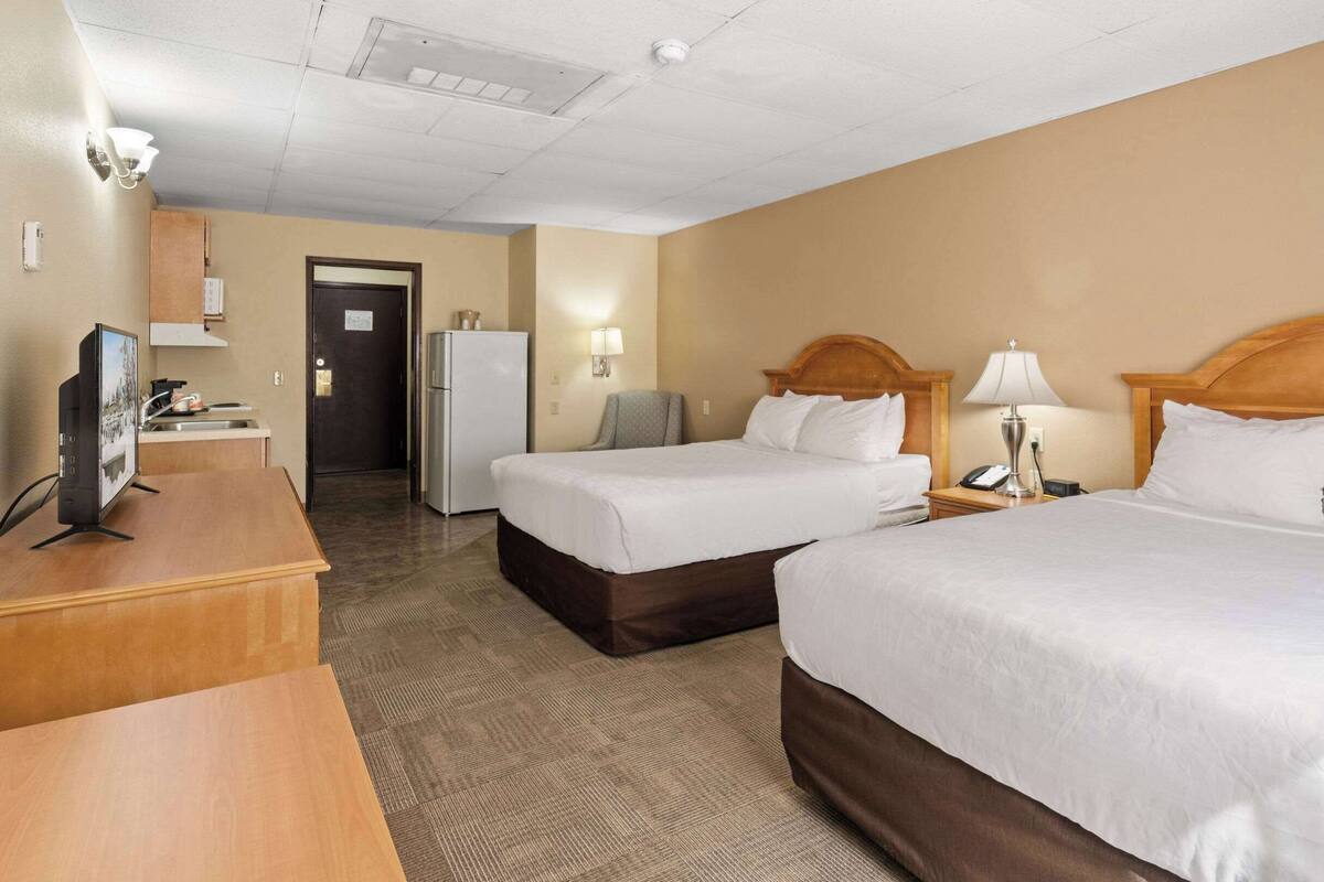 A hotel room with two beds, a desk with a computer, a refrigerator, a lamp, and a door leading to a bathroom.