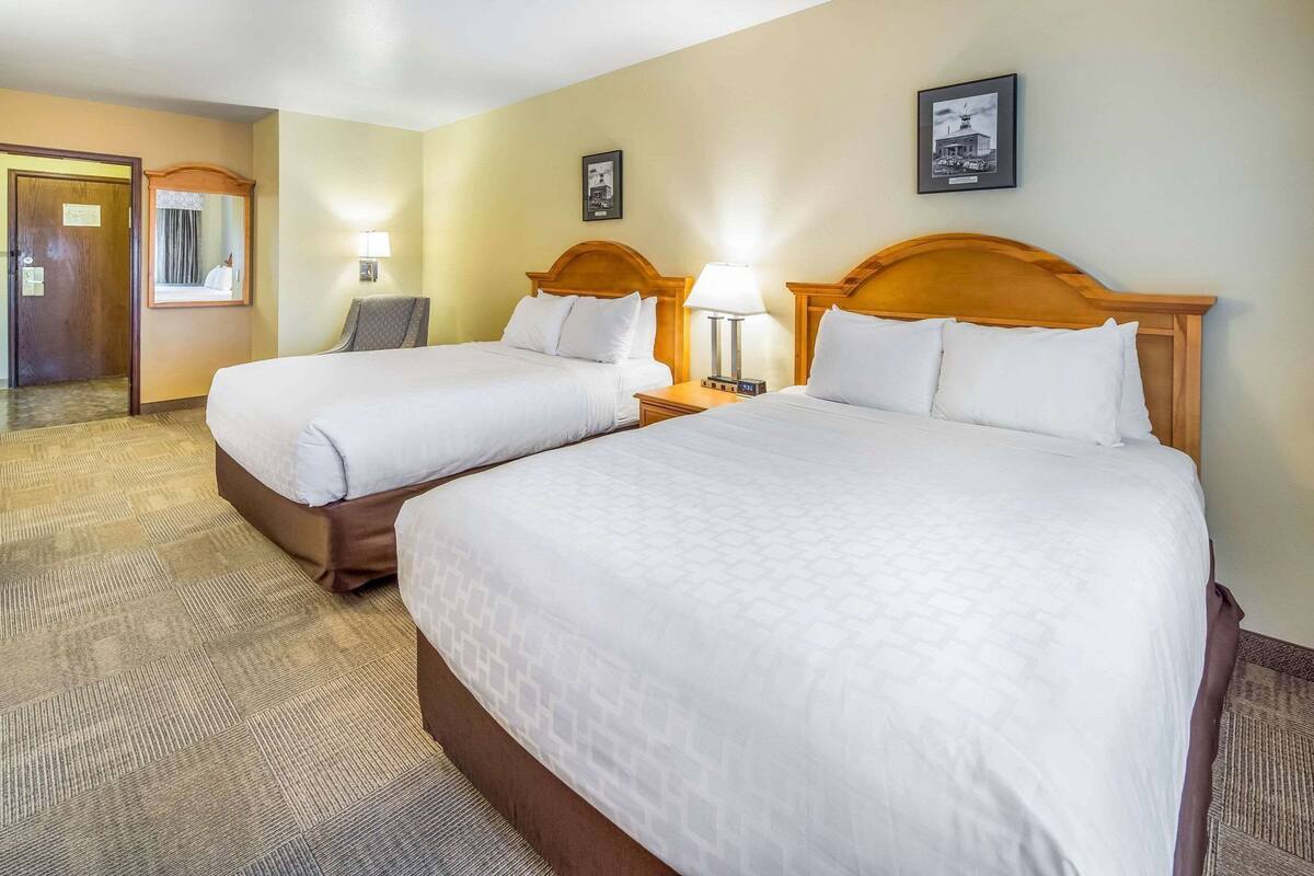 A hotel room with two beds, a wooden headboard, a nightstand with a lamp, a mirror, and a framed picture on the wall.