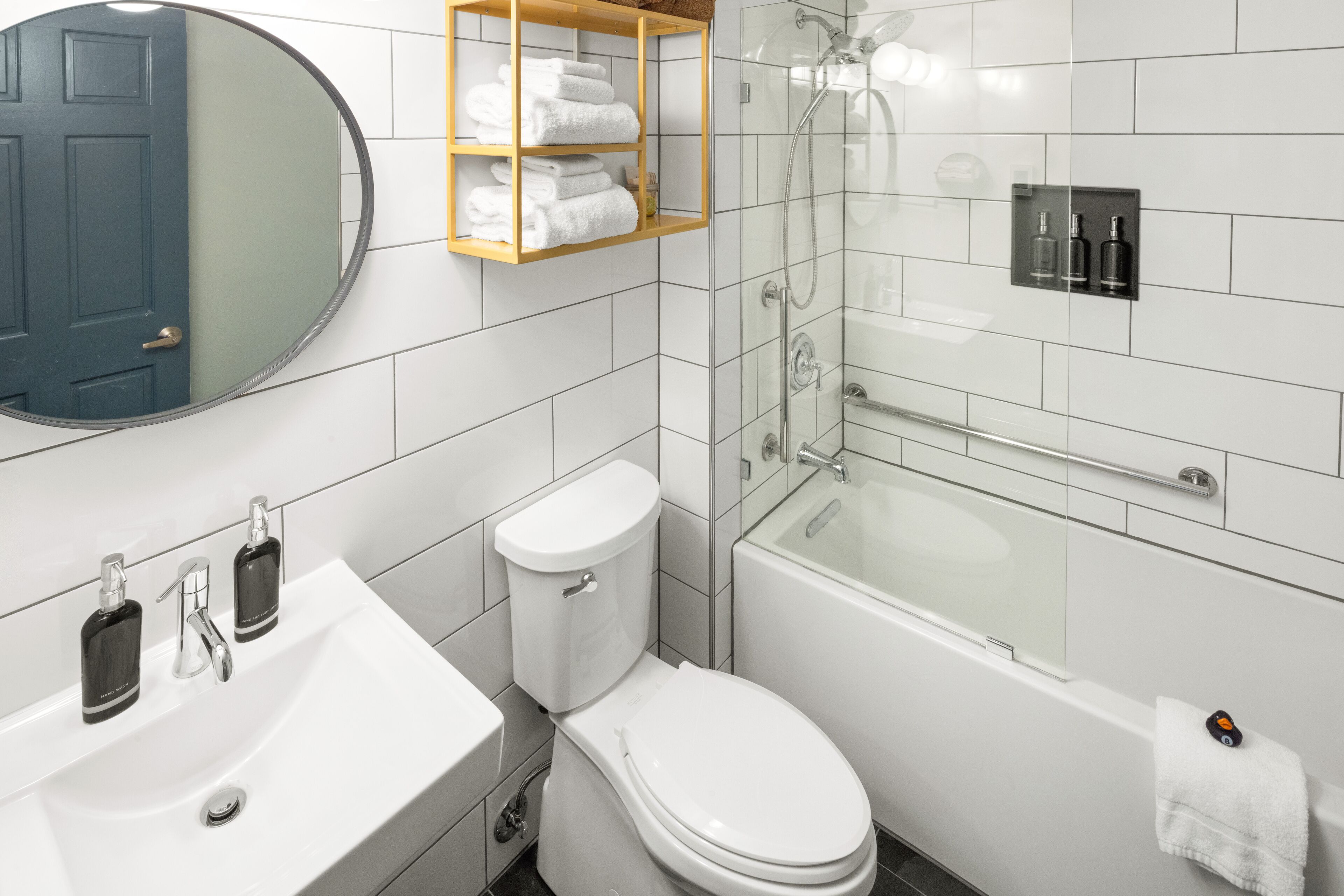 A modern bathroom with a white sink, toilet, and bathtub. There is a glass shower enclosure and a wooden shelf with towels.