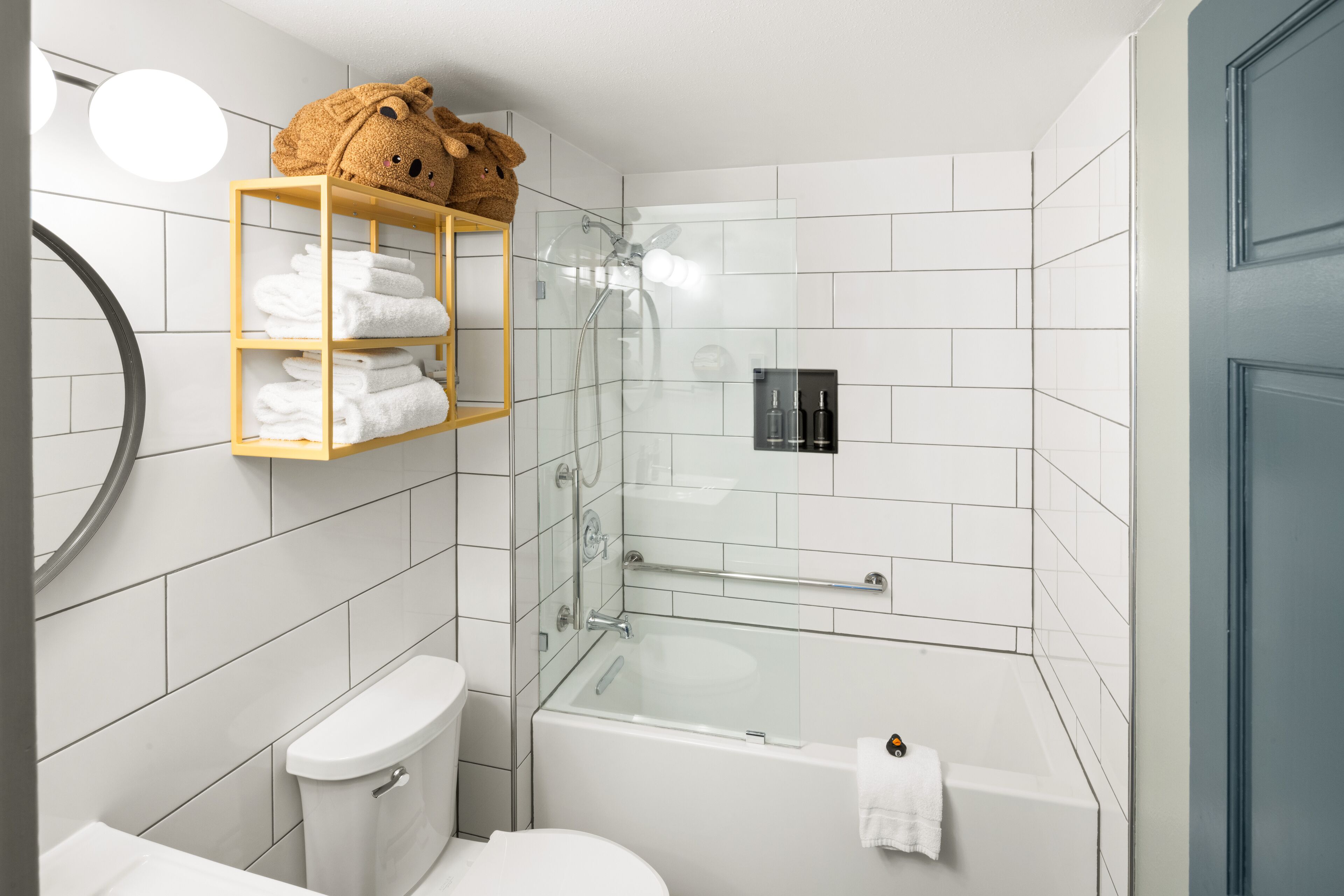 A modern bathroom with a glass shower enclosure, a white bathtub, and a toilet. There is a wooden shelf with towels and a teddy bear.