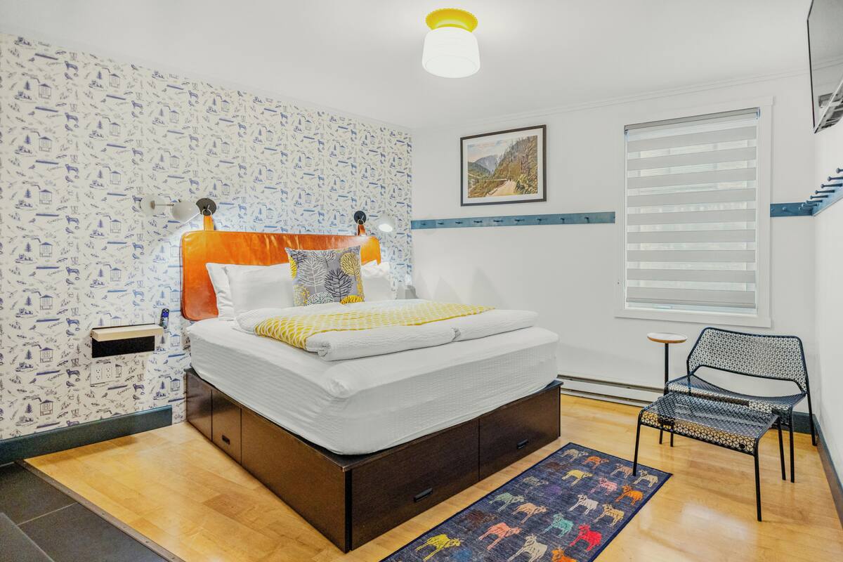 A neatly arranged bedroom with a bed, a chair, a window with blinds, and a patterned rug.