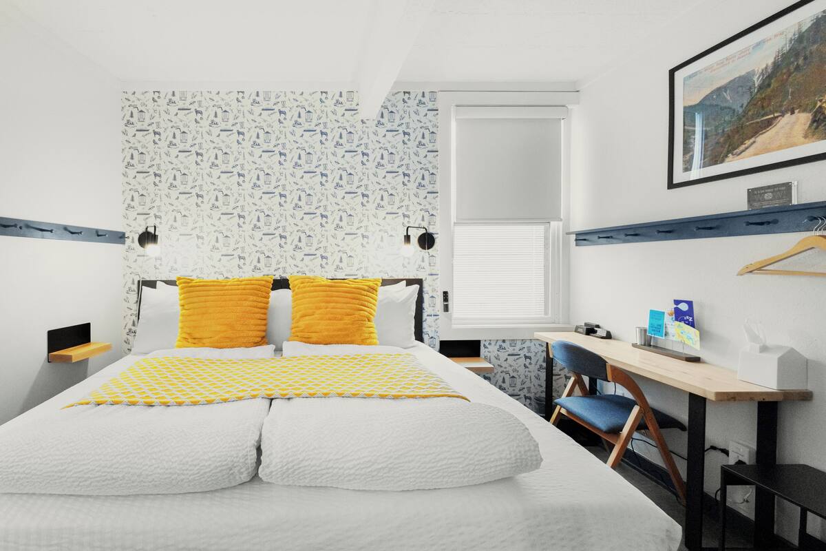 A bedroom with a bed, a desk, a chair, a window, and a wall-mounted shelf.