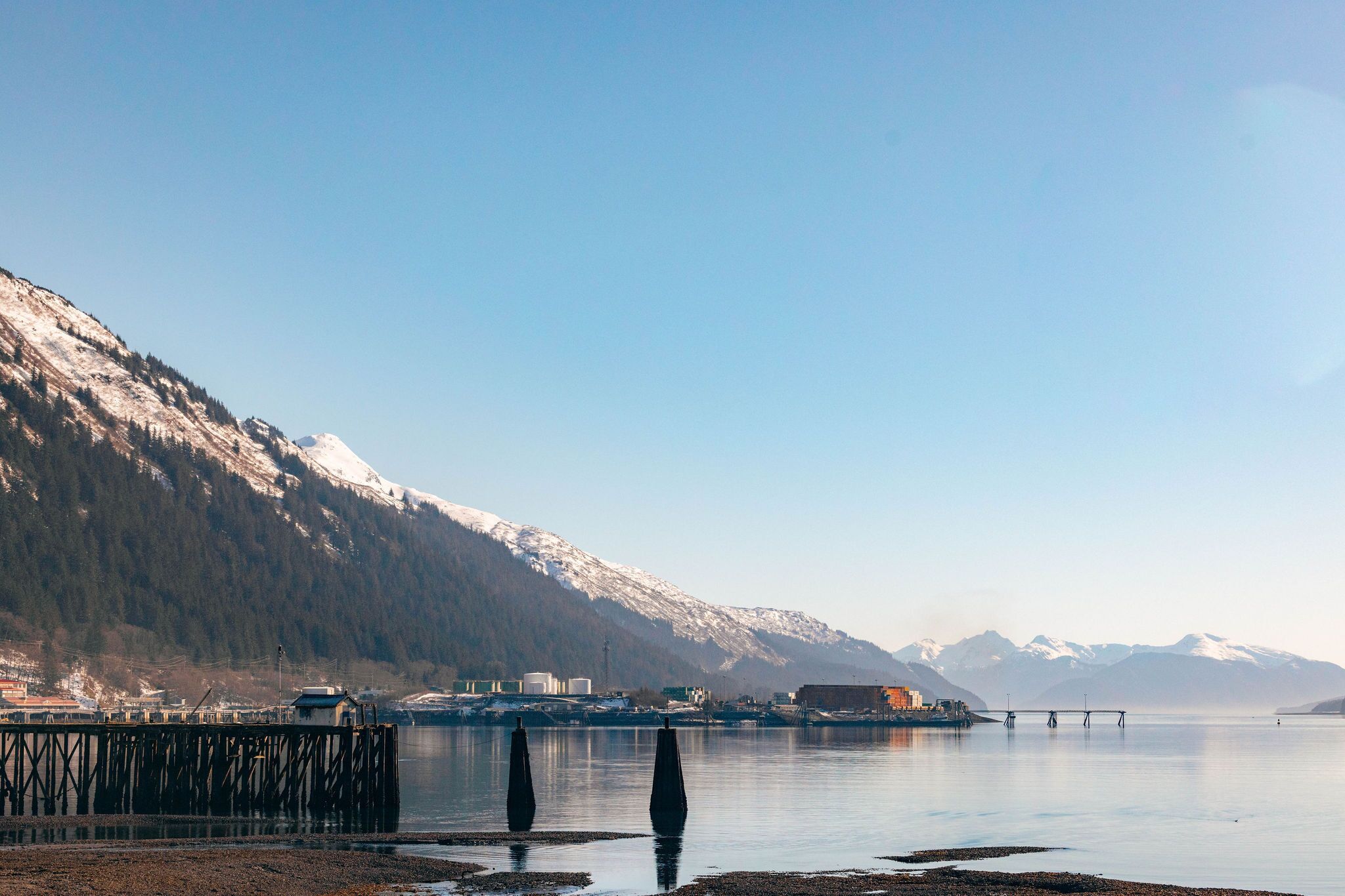Een rustig meer met een houten steiger die het water in loopt, omgeven door bergen met besneeuwde toppen.