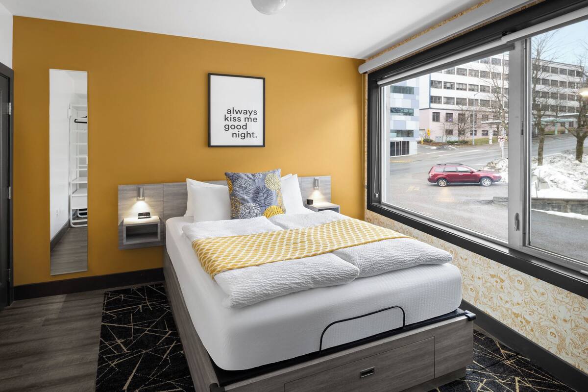 A hotel room with a bed, bedside tables, a mirror, a window with a view of the street, and a motivational quote on the wall.