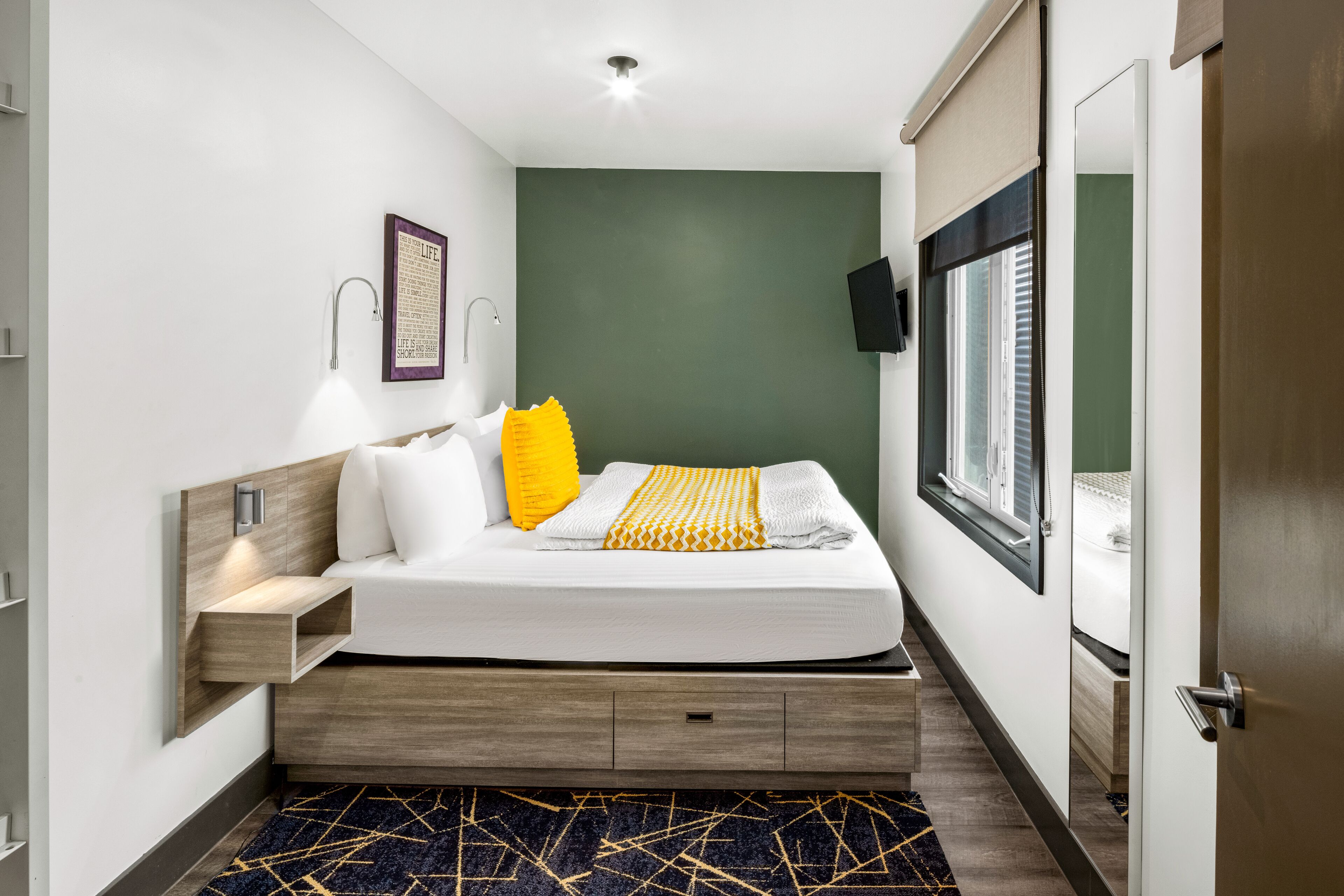 A modern hotel room with a bed, bedside table, wall-mounted TV, and a window with blinds.