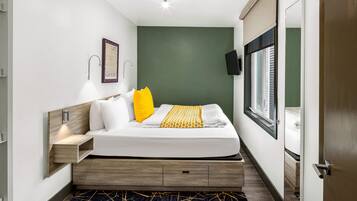 A modern hotel room with a bed, bedside table, wall-mounted TV, and a window with blinds.