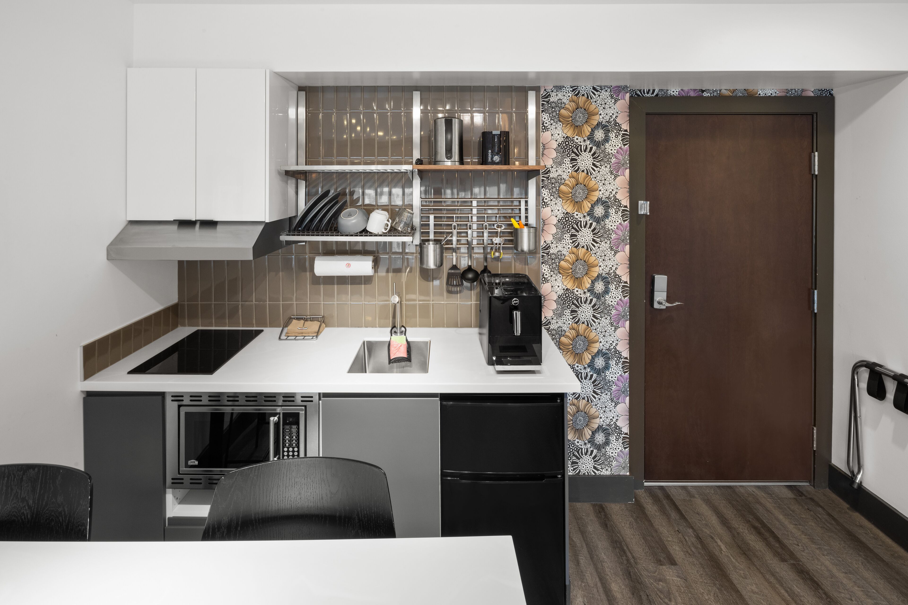 A compact kitchen with a microwave, dishwasher, and a coffee maker. There is a sink with a faucet, and a wooden shelf above it. The kitchen has a white countertop and a patterned backsplash.