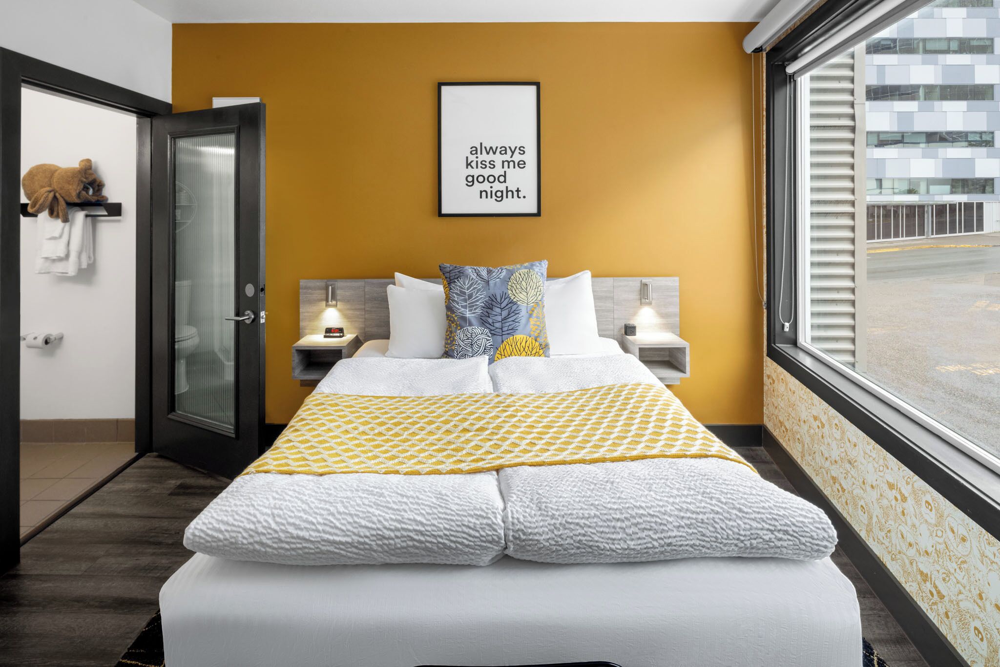 A hotel room with a bed, a bedside table, a nightstand, a window with a view of the street, and a wall with a framed quote.