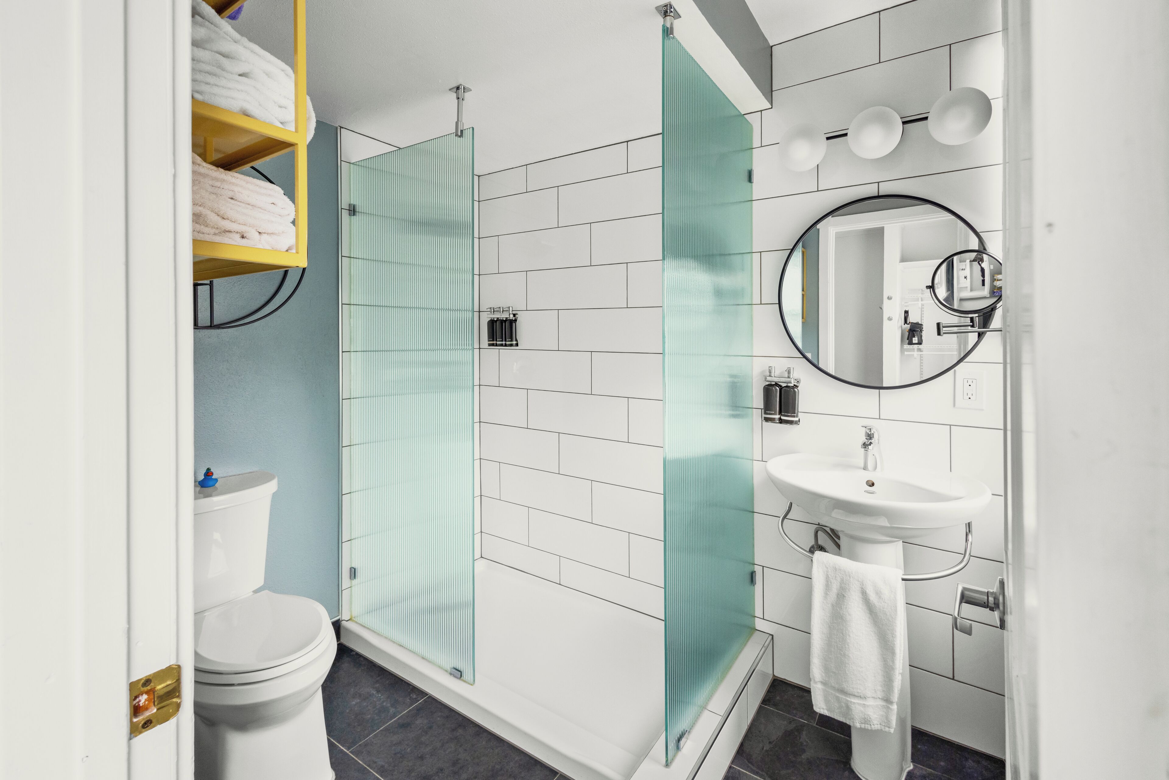 A modern bathroom with a glass shower enclosure, white tiles, a white sink, and a mirror.