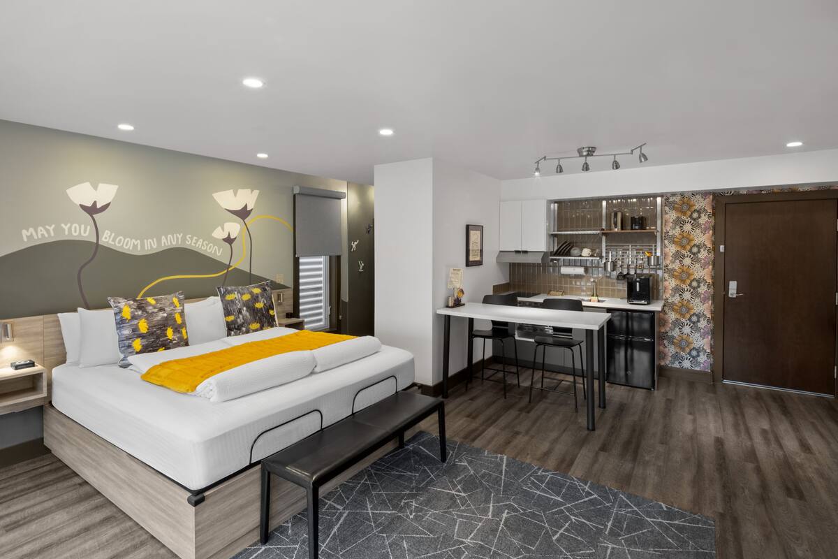 A modern bedroom with a bed, bedside table, and a bench. It includes a small kitchenette with a fridge, sink, and bar stools.