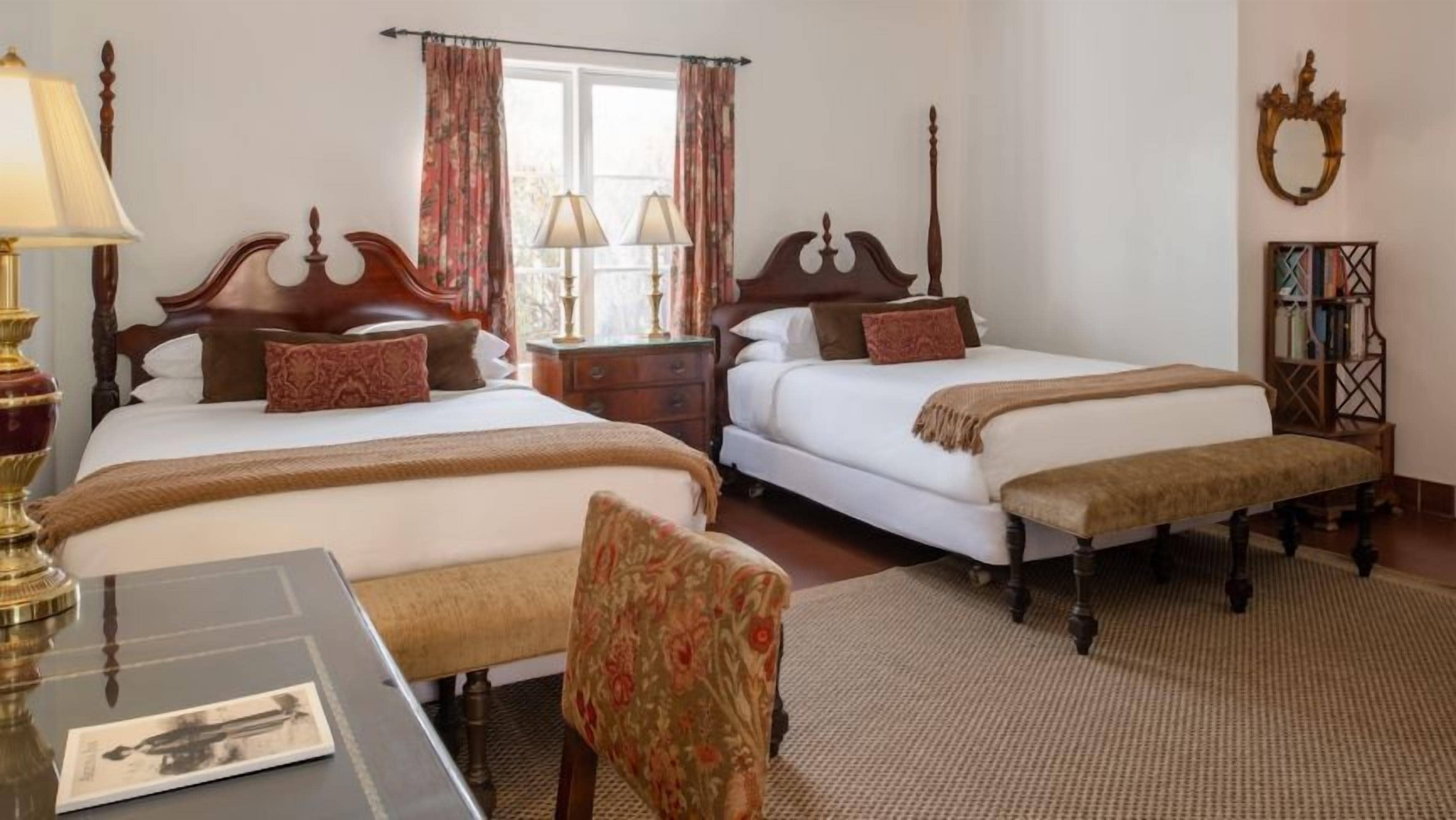 A hotel room with two beds, a desk, a chair, and a bookshelf.