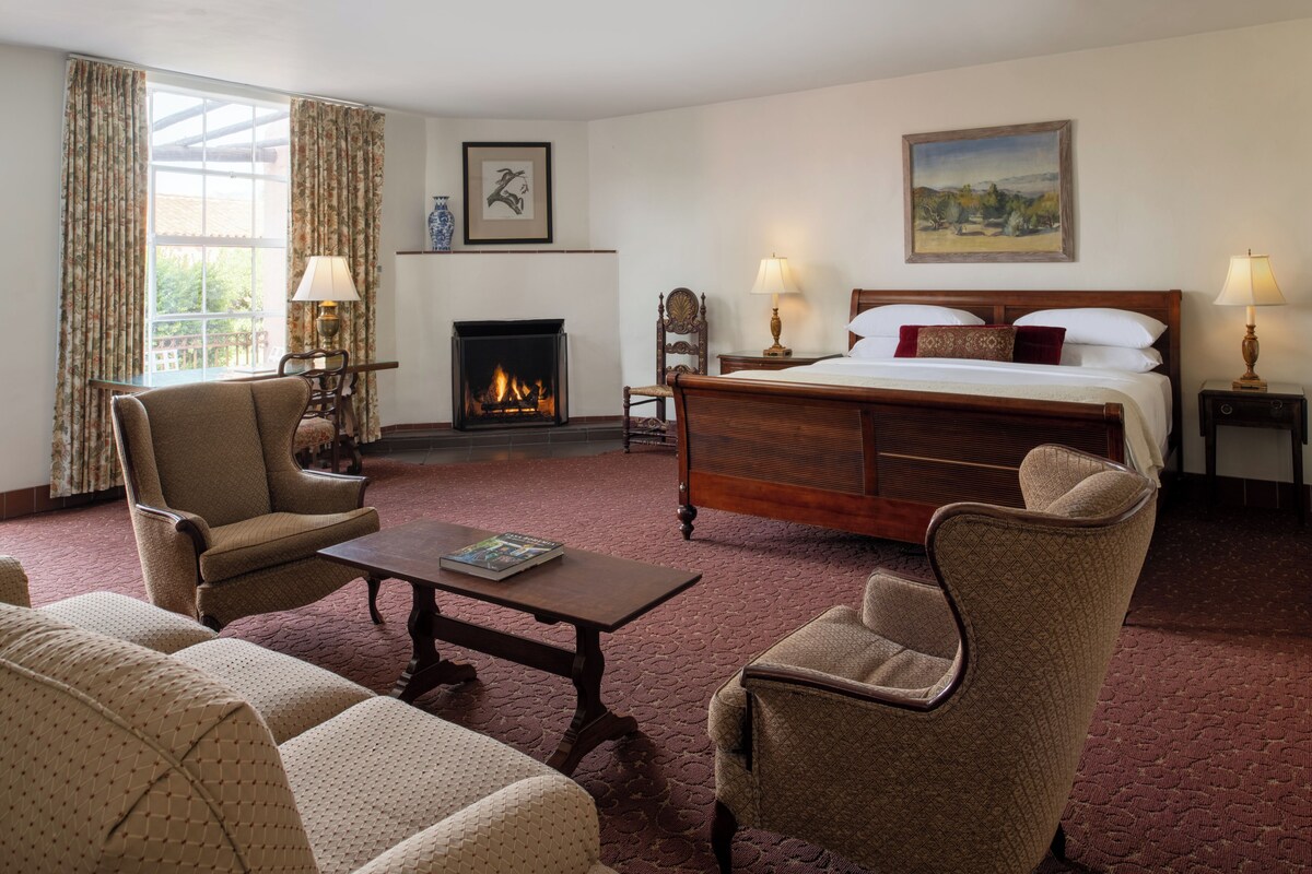 Een hotelkamer met een groot bed, twee fauteuils, een houten salontafel, een open haard en een schilderij aan de muur.