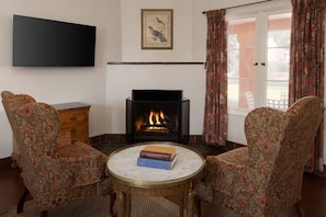 Una sala de estar con chimenea, dos sillones, una mesa de centro con libros, un televisor y un cuadro en la pared.