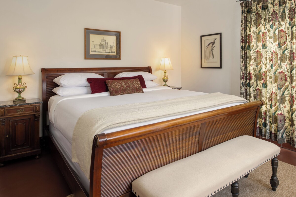 Een net opgemaakt bed met een houten hoofdbord, twee bedlampjes en een bank aan het voeteneinde.