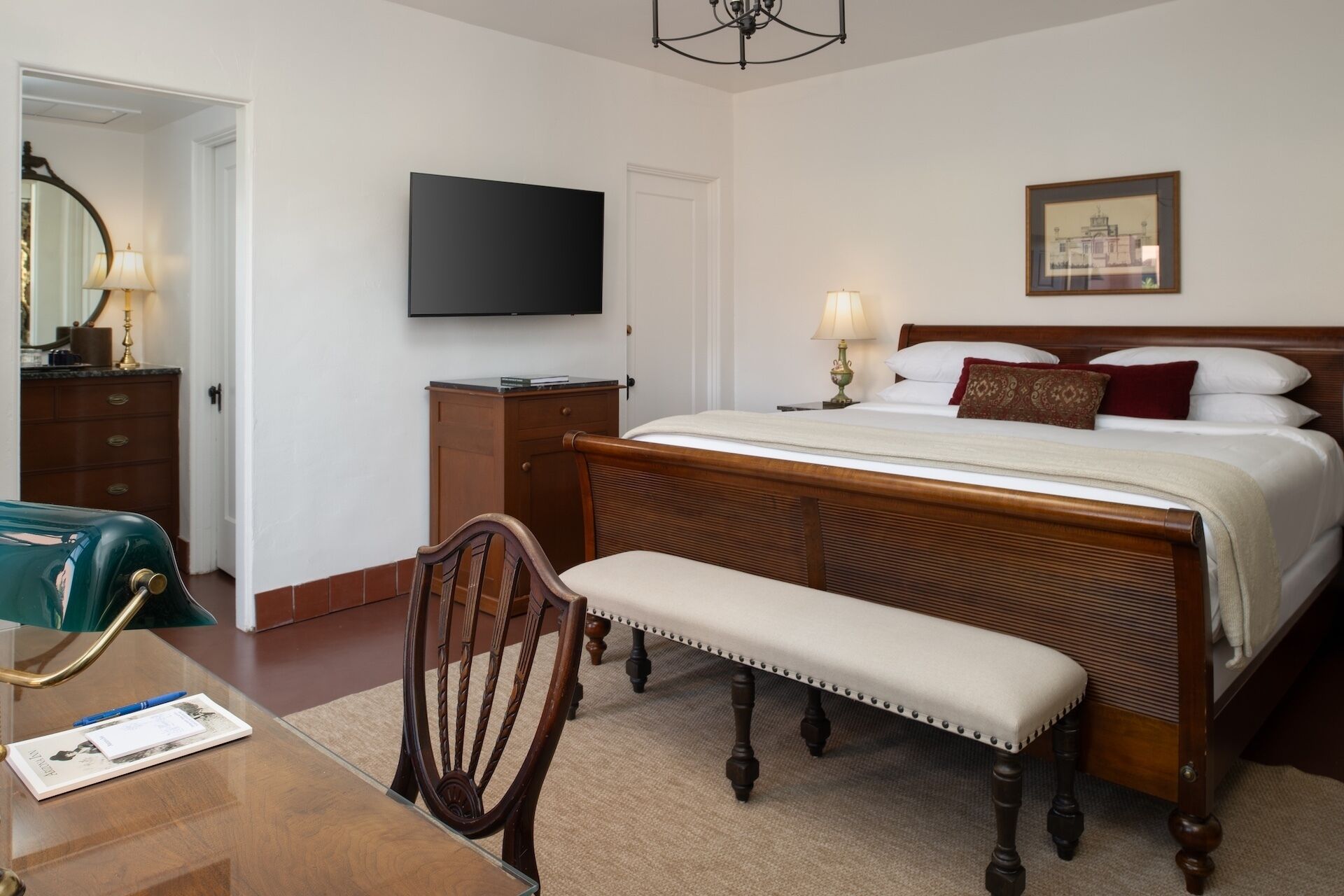 A hotel room with a bed, bedside table, chair, desk, TV, and a framed picture on the wall.
