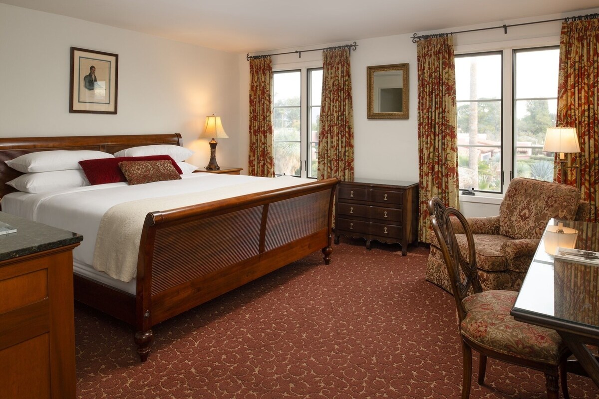Een hotelkamer met een houten bed, een bureau met stoel, een dressoir en een raam met gordijnen.