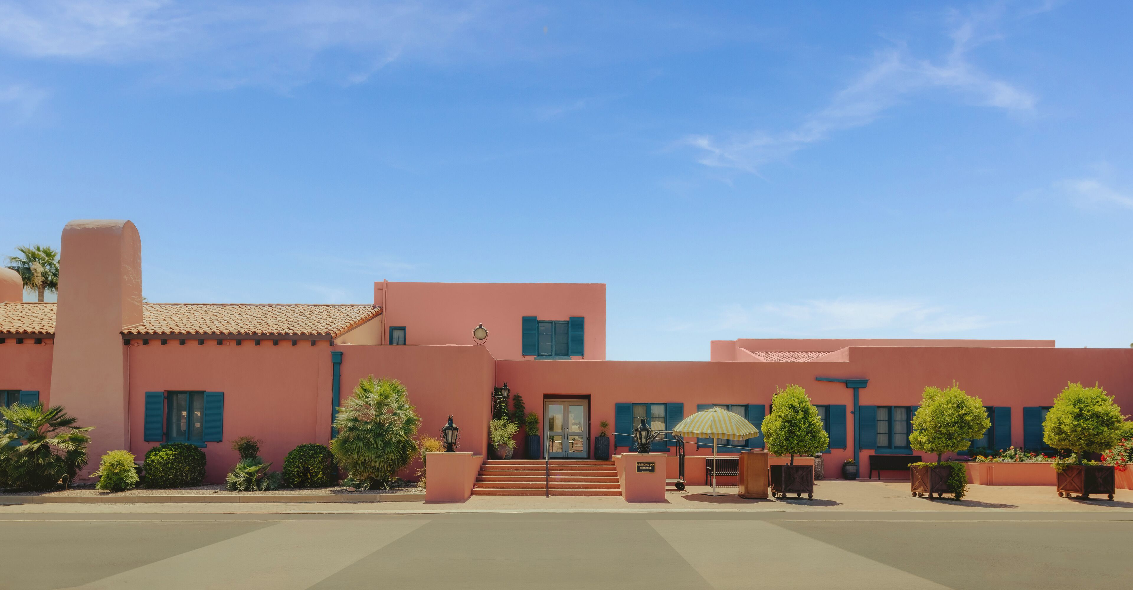 A pink building with a red-tiled roof, a set of stairs leading to the entrance, and a small outdoor seating area with a striped umbrella.