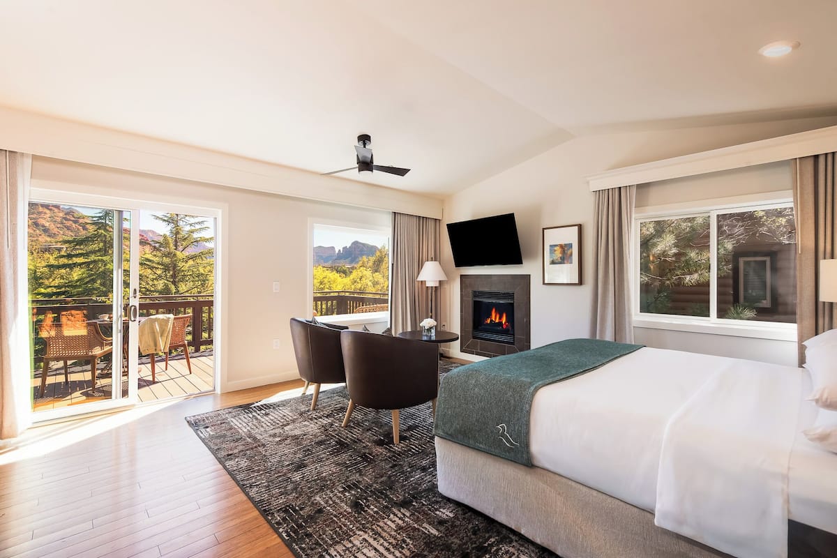 Habitación de hotel con cama, escritorio con sillas, chimenea, televisor y balcón con vista a árboles y montañas.