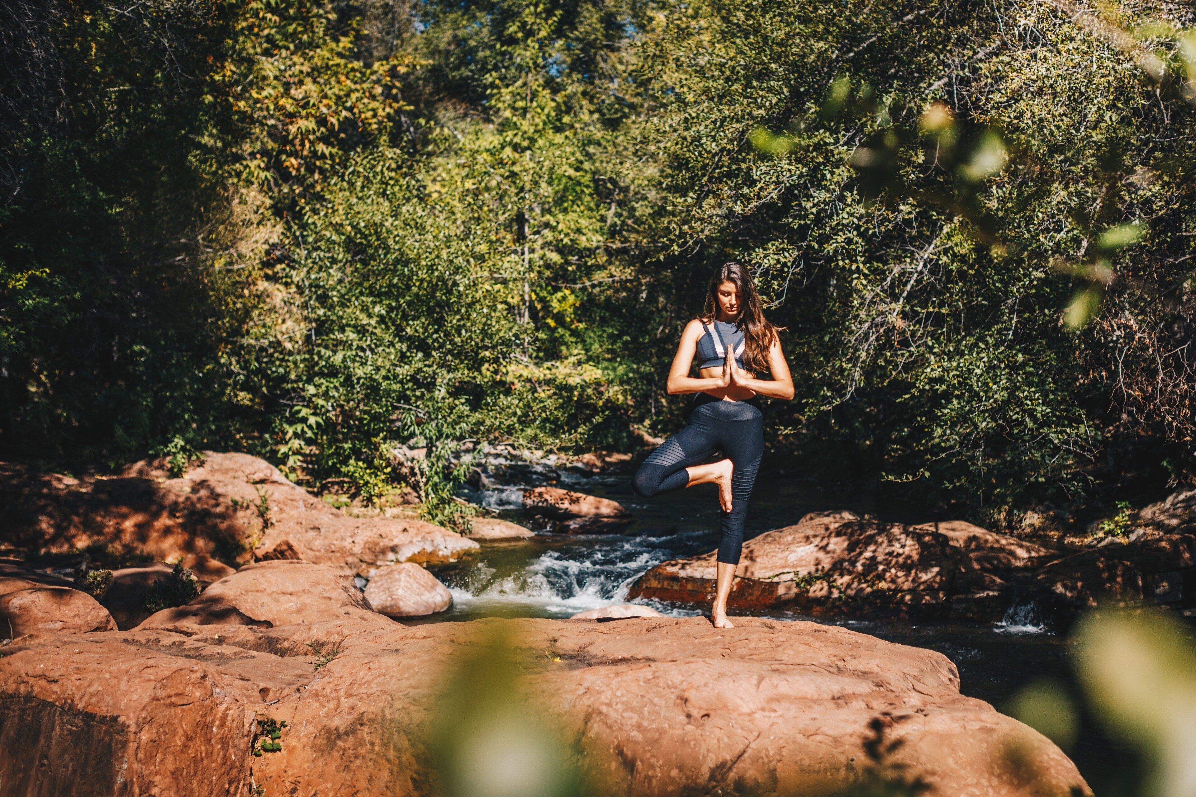 A person performing a yoga pose on a rock by a stream in a forested area.