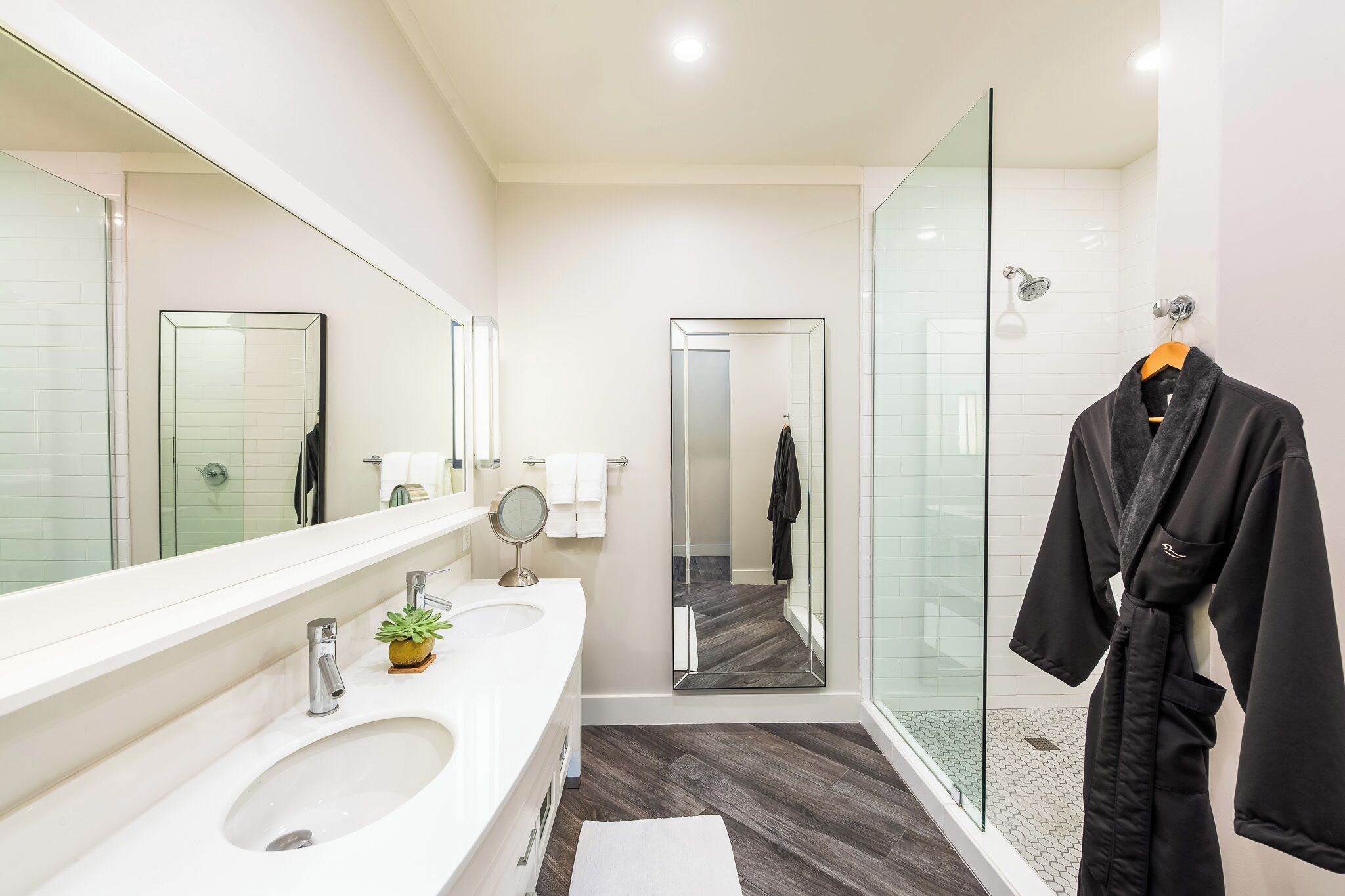 A modern bathroom with a large mirror, two sinks, a shower, and a robe hanging on a rack.