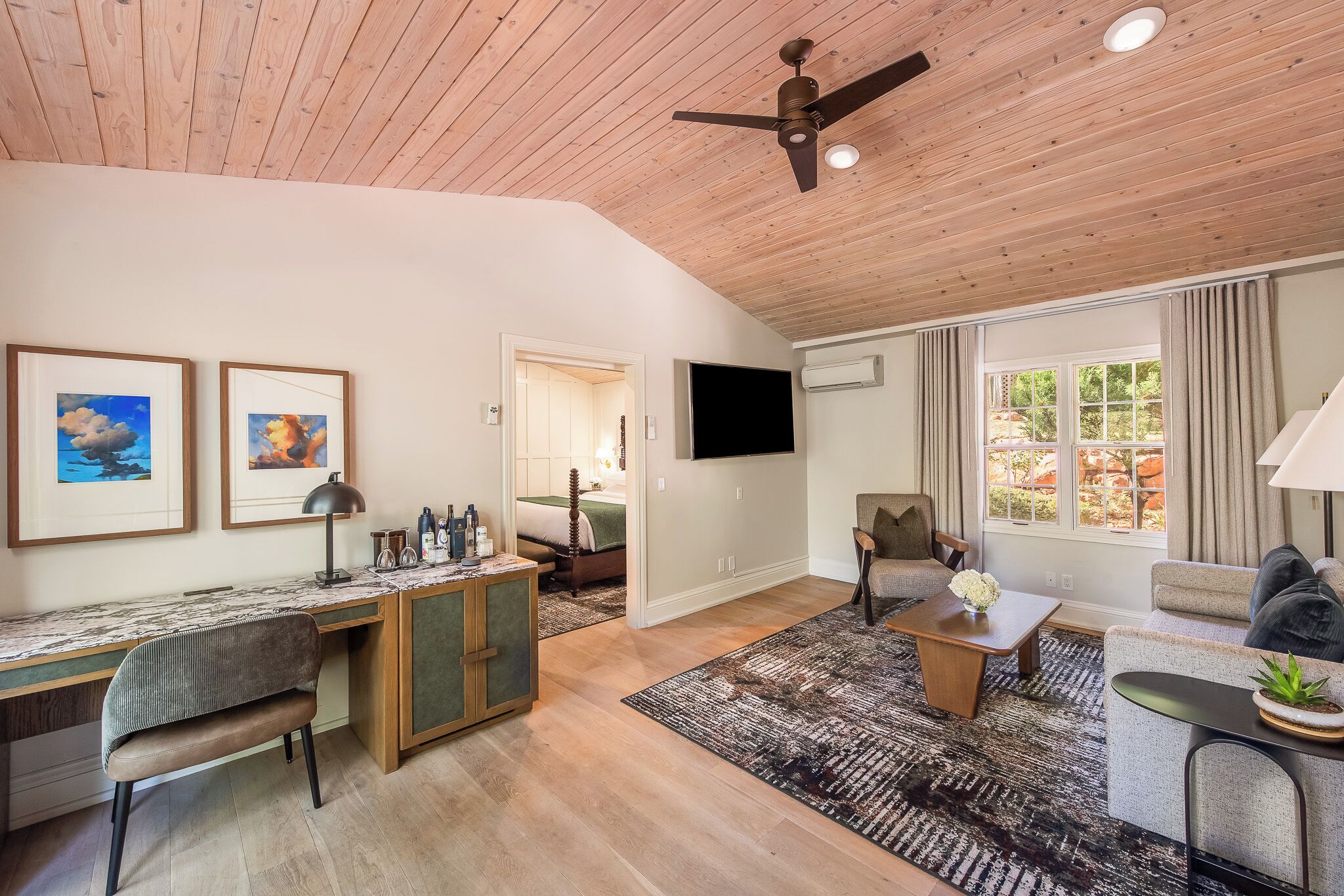 A spacious living area with a wooden ceiling, a flat-screen TV, a desk with a chair, a sofa, and a coffee table.