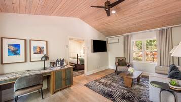 A spacious living area with a wooden ceiling, a flat-screen TV, a desk with a chair, a sofa, and a coffee table.