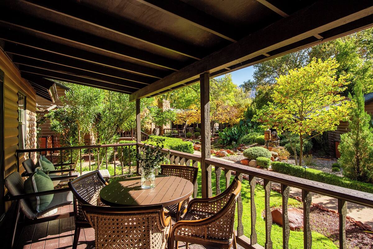 Un patio exterior techado con una mesa redonda y sillas de mimbre, rodeado por un jardín exuberante con diversas plantas y árboles.