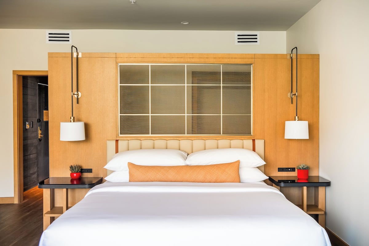 Habitación de hotel con una cama grande, cabecera de madera, dos mesitas de noche con lámparas y una ventana con persianas.