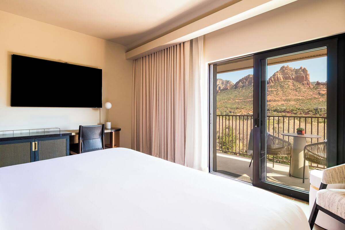 Habitación de hotel con cama, escritorio, silla, televisor y balcón con vistas a terreno rocoso.