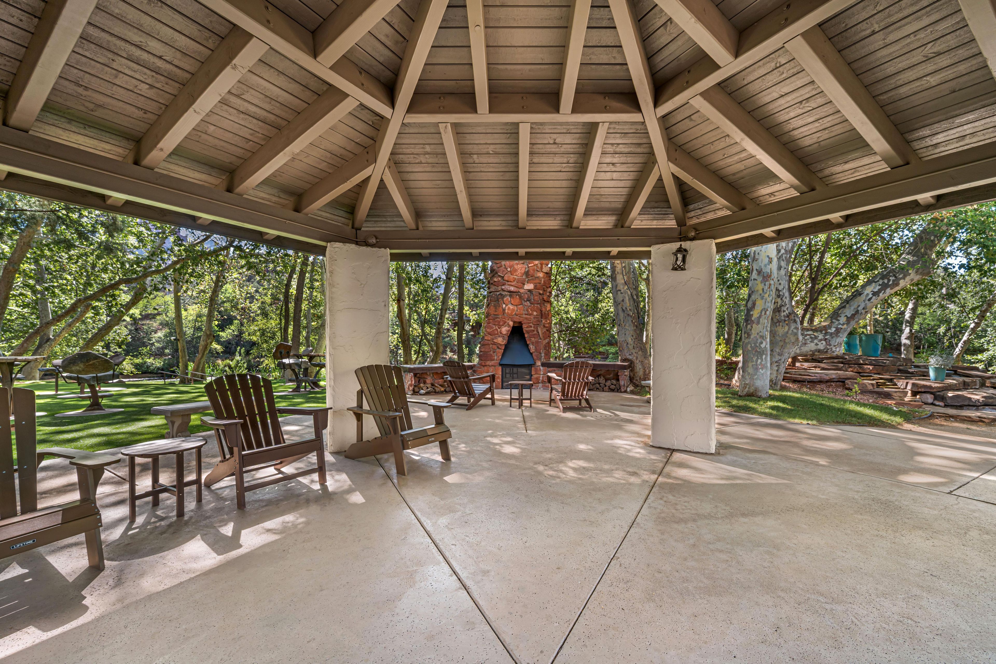 A covered outdoor area with Adirondack chairs, a stone fireplace, and a view of the surrounding trees.