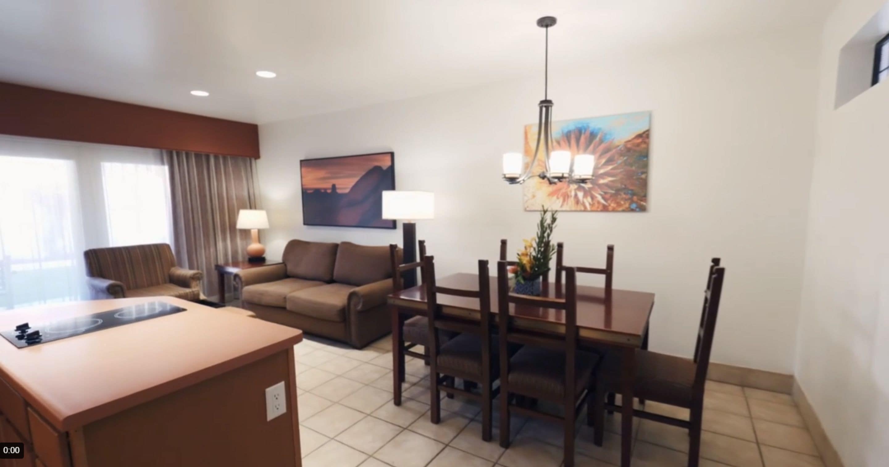 A hotel room with a kitchenette, a dining area with a table and chairs, a sofa, a lamp, and a painting on the wall.