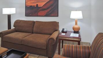 A brown sofa, a striped armchair, a small wooden table with a lamp, and a painting of a sunset on the wall.