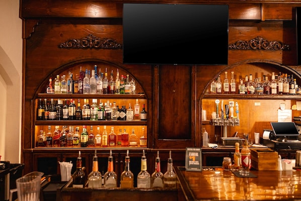 Zona de bar con estantes llenos de botellas de licor, un televisor de pantalla plana y una caja registradora.