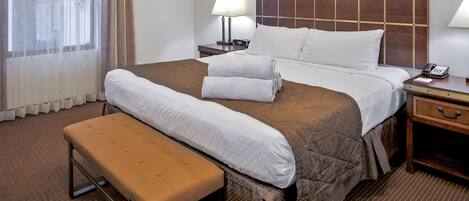 Hotellihuone, jossa on suuri sänky, yöpöytä lampun kanssa, penkki ja ikkuna, jossa on verhot.