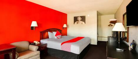 A hotel room with a red accent wall, a bed with a white and red bedspread, a striped armchair, a desk with a lamp, and a television.