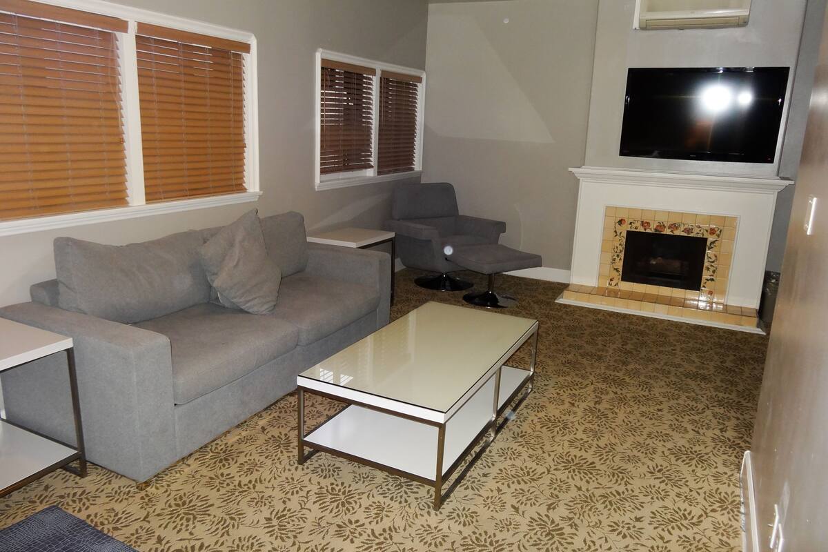 A living room with a grey sofa, a small white table, a TV mounted above a fireplace, and patterned carpet.