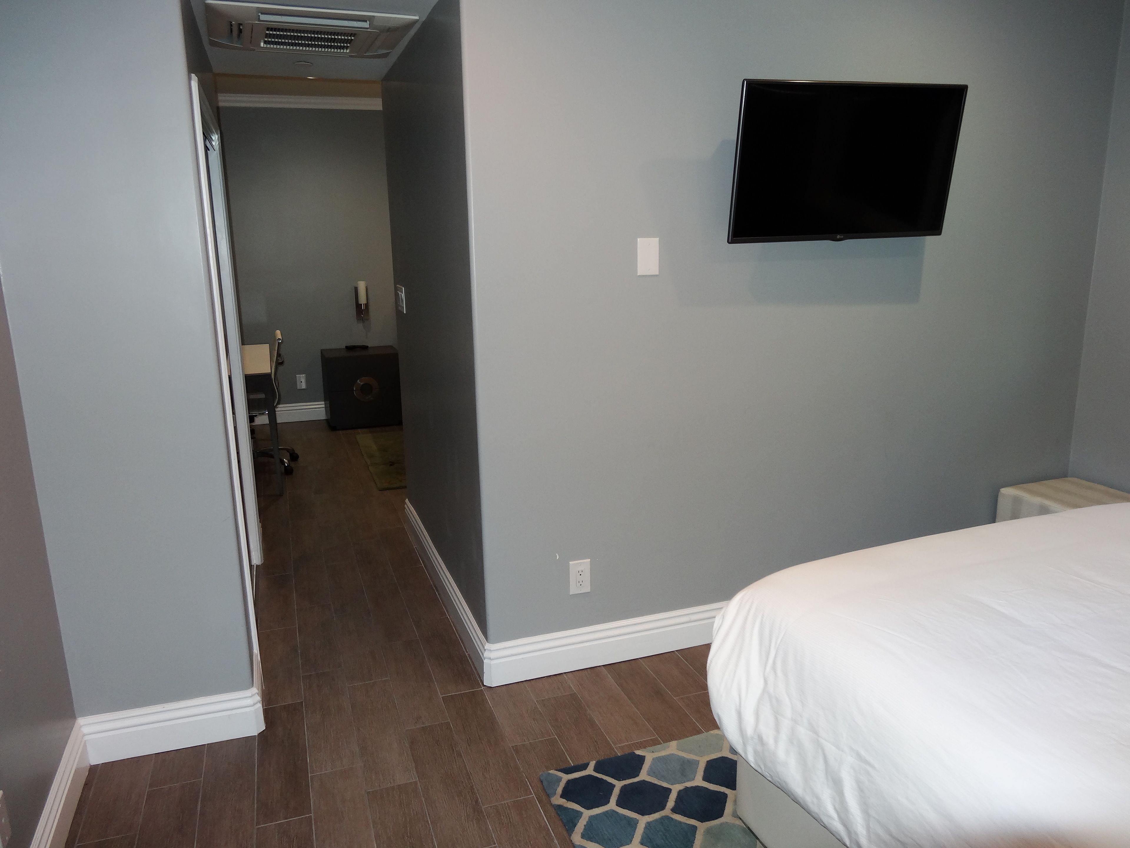 A bedroom with a bed, a TV mounted on the wall, a small bedside table, and a door leading to another room.