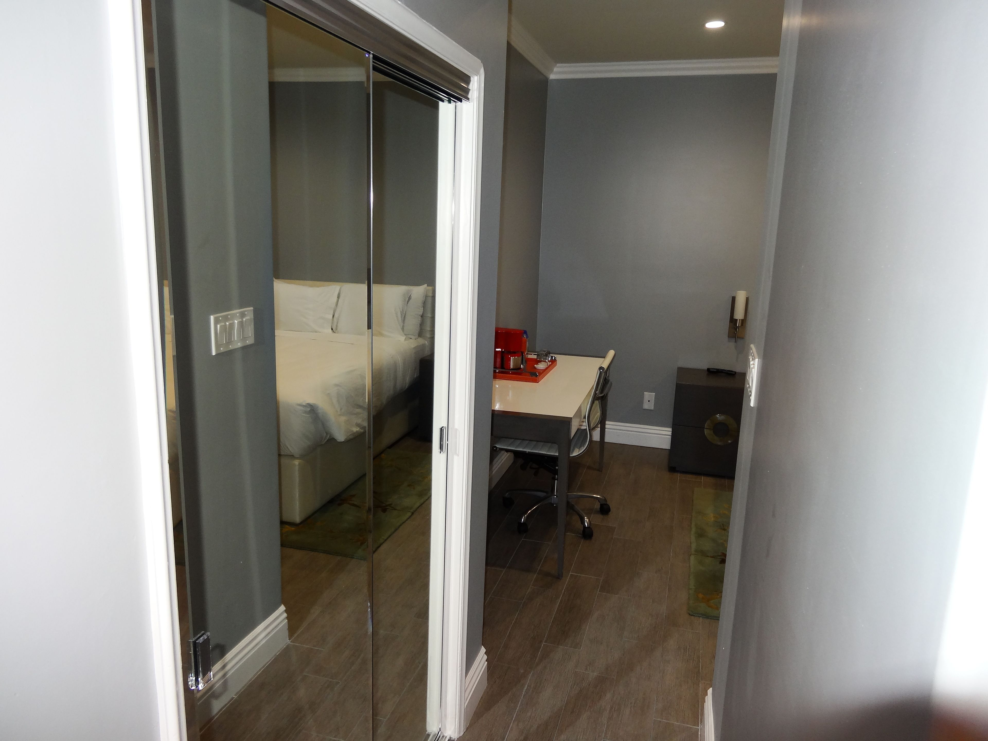 Une chambre à coucher avec un lit, un bureau et une chaise. Il y a une fenêtre avec des stores, une porte et une petite table sur laquelle se trouve un objet rouge.