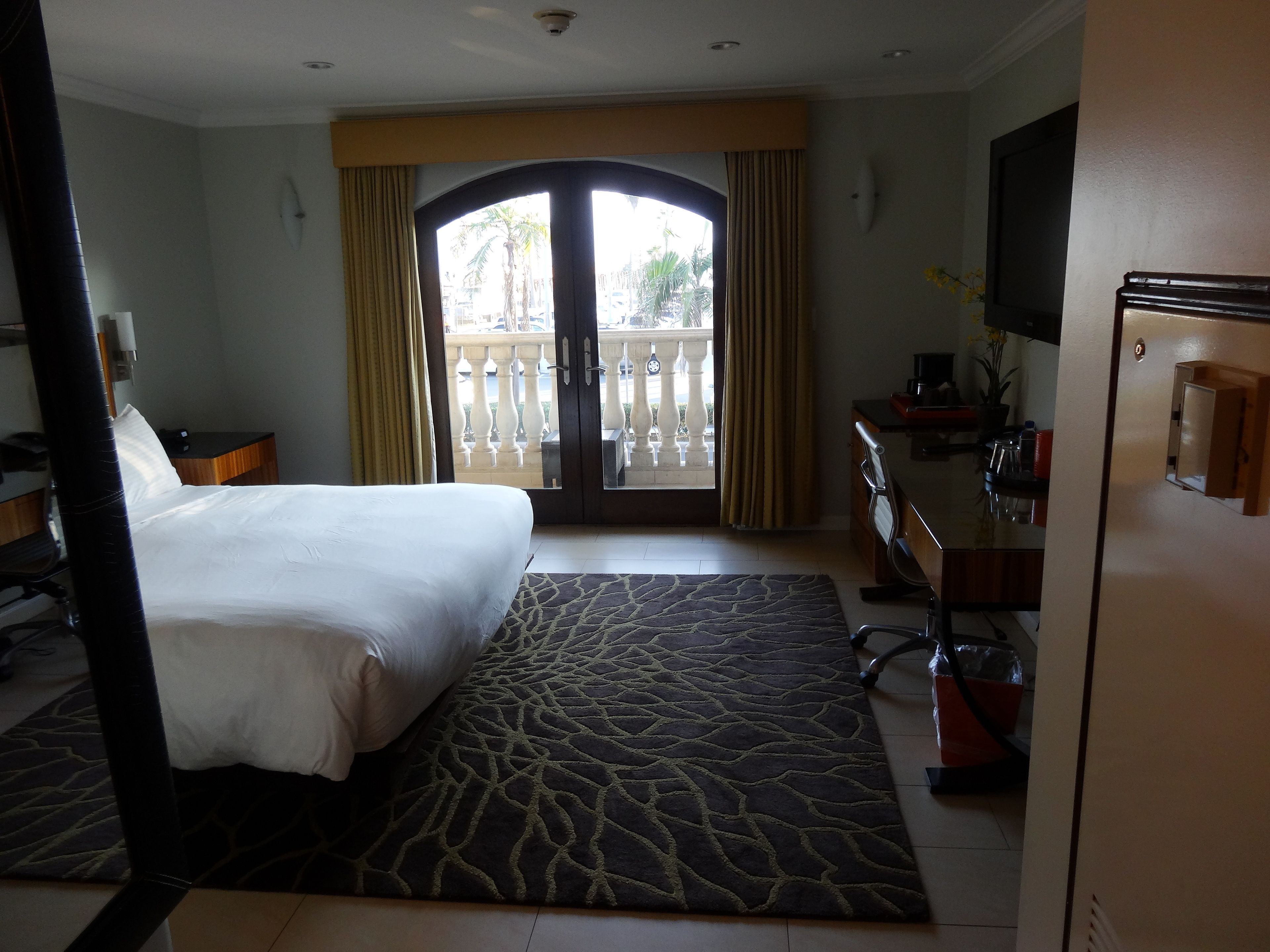 A hotel room with a bed, desk, chair, and a view of the outside balcony.