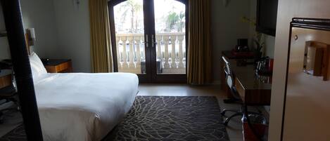 A hotel room with a bed, desk, chair, and a view of the outside balcony.