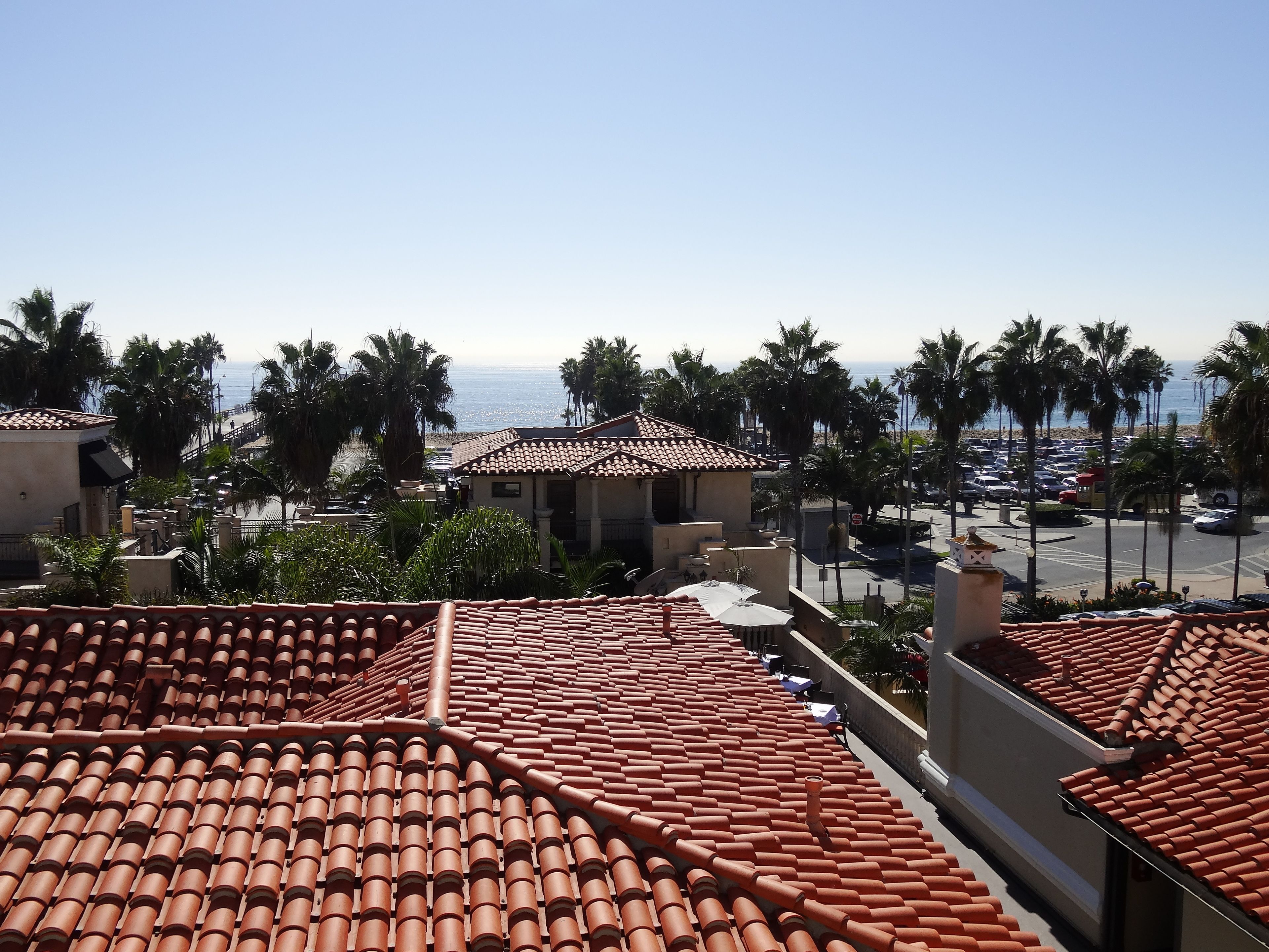 A coastal view with terracotta tiled roofs, palm trees, and a parking lot.