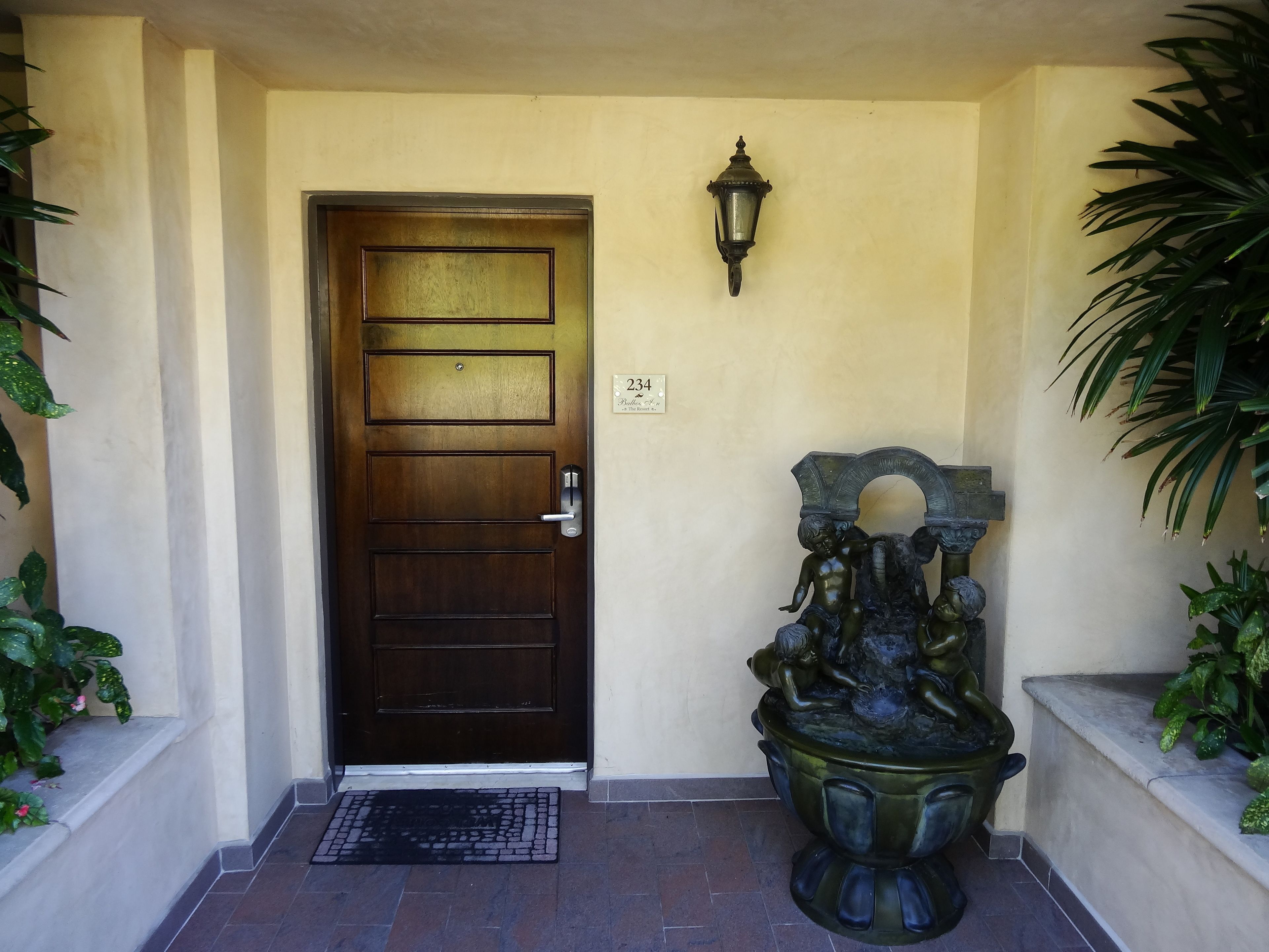 A wooden door with a metal handle and a doormat. There is a wall-mounted light fixture above the door and a decorative sculpture to the right.
