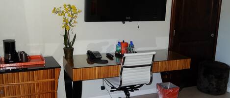 Une chambre d’hôtel avec un bureau en bois, une chaise noire, une petite plante en pot, une machine à café, un téléphone et un téléviseur à écran plat fixé au mur.
