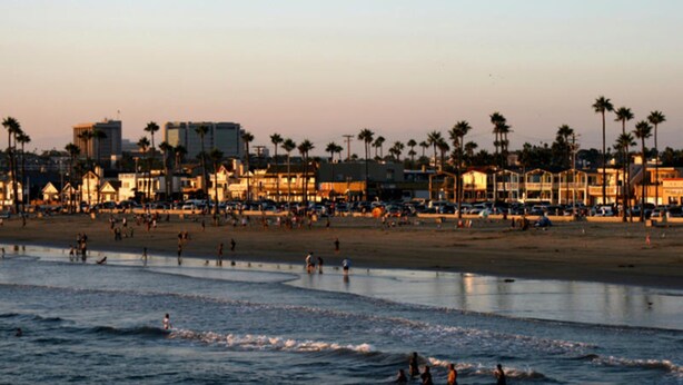 A beach with people swimming and walking along the shore. There are palm trees and buildings in the background. - Balboa Inn, On the Beach (Newport Beach)