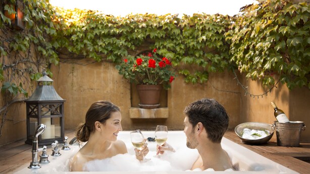 Una pareja en un jacuzzi, con una planta en maceta y una vela al fondo.