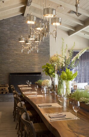 A long wooden dining table set for a meal with chairs, a chandelier, and a fireplace in the background.