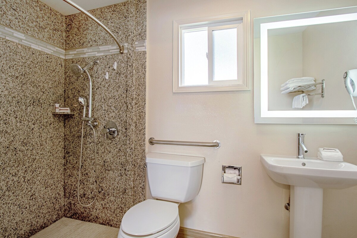 A bathroom with a granite shower wall, a white toilet, a sink with a mirror above it, and a window providing natural light.