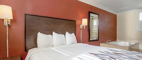 Quarto de hotel com cama grande, mesa de cabeceira, banheiro com pia e espelho e luminária na parede.