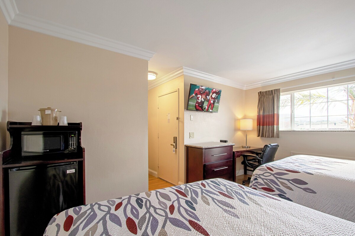 A hotel room with two beds, a desk, a chair, a microwave, a refrigerator, a television, and a window with curtains.